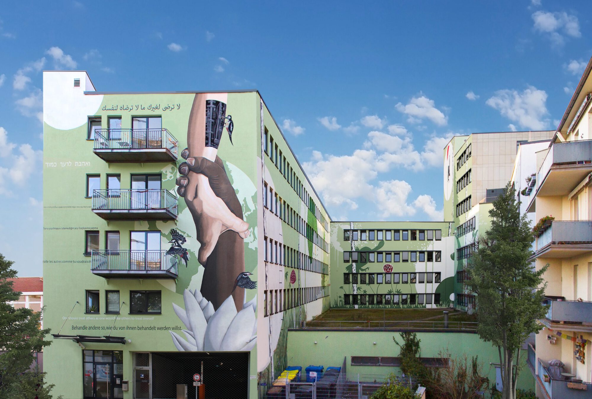 Fassadengestaltung eines Gebäudes mit einem großen Wandgemälde, das zwei ineinander greifende Hände darstellt. Auf dem grünen Gebäude sind bunte Blumen und Vögel abgebildet, dazu vielsprachige Zitate über Mitmenschlichkeit. Konzeptionelle Kunst von Goodlack Art im urbanen Kontext.