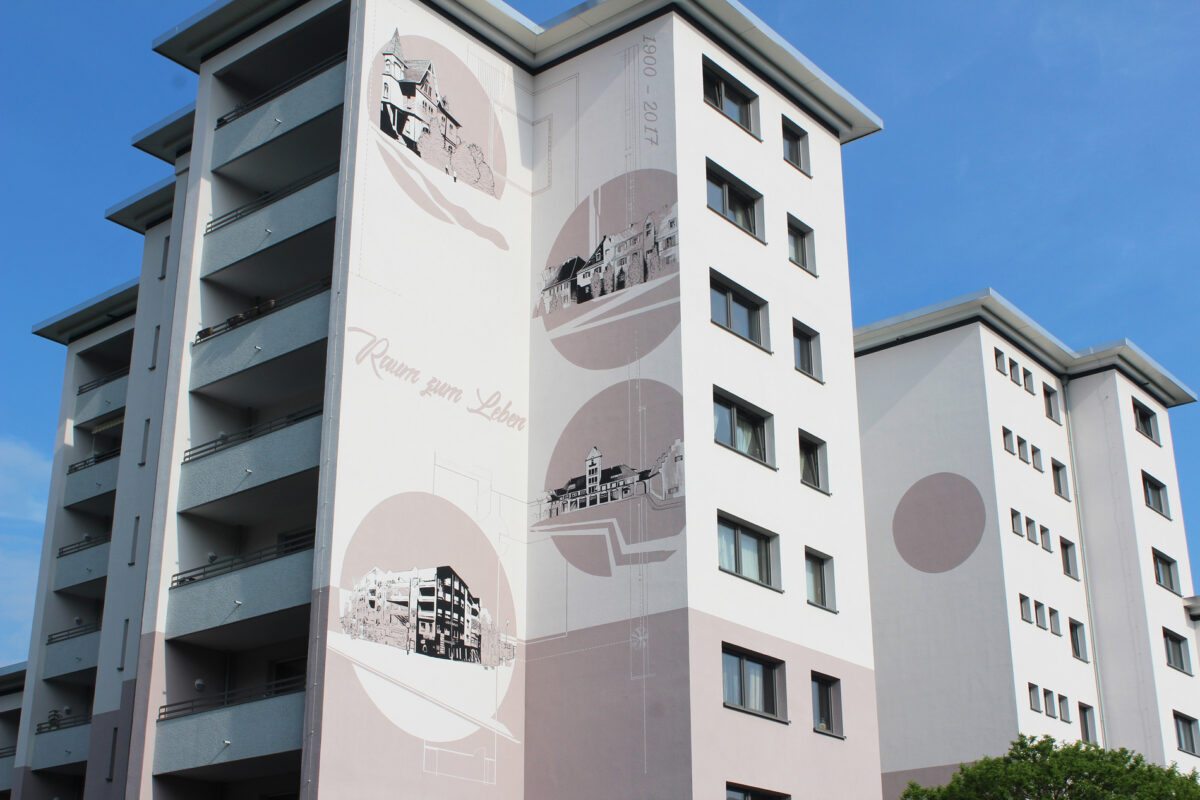 Skyscrapers in Cologne featuring large-scale facade art by Goodlack Art, depicting stylized architecture and the words "Raum zum Leben" (Space to Live), under a blue sky.