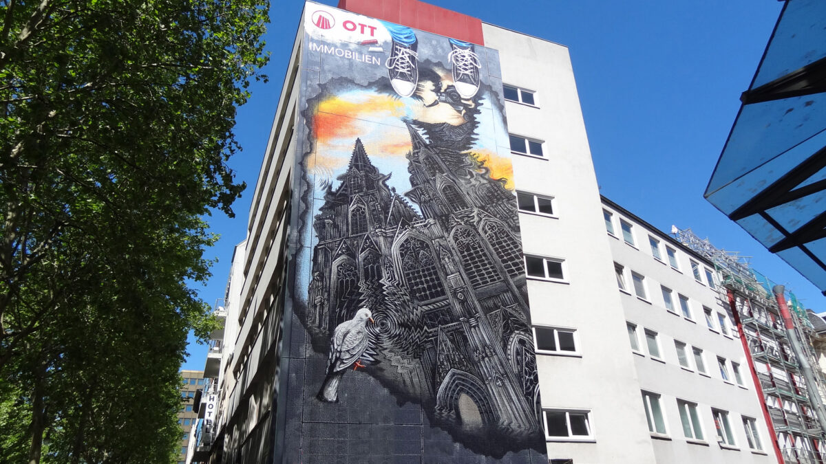 A large mural on a building in Cologne features a creative depiction of architectural elements and a dove. At the top, painted sneakers reach up toward the sky, while the background suggests a colorful sunset. The artwork is an example of street art in the city.
