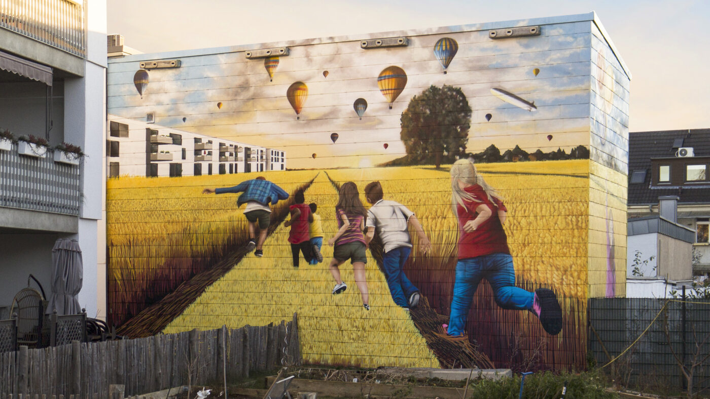 Wandgemälde in Köln von Goodlack Art zeigt Kinder, die durch ein Weizenfeld rennen, mit Heißluftballons und einem Zeppelin im Himmel im Hintergrund.