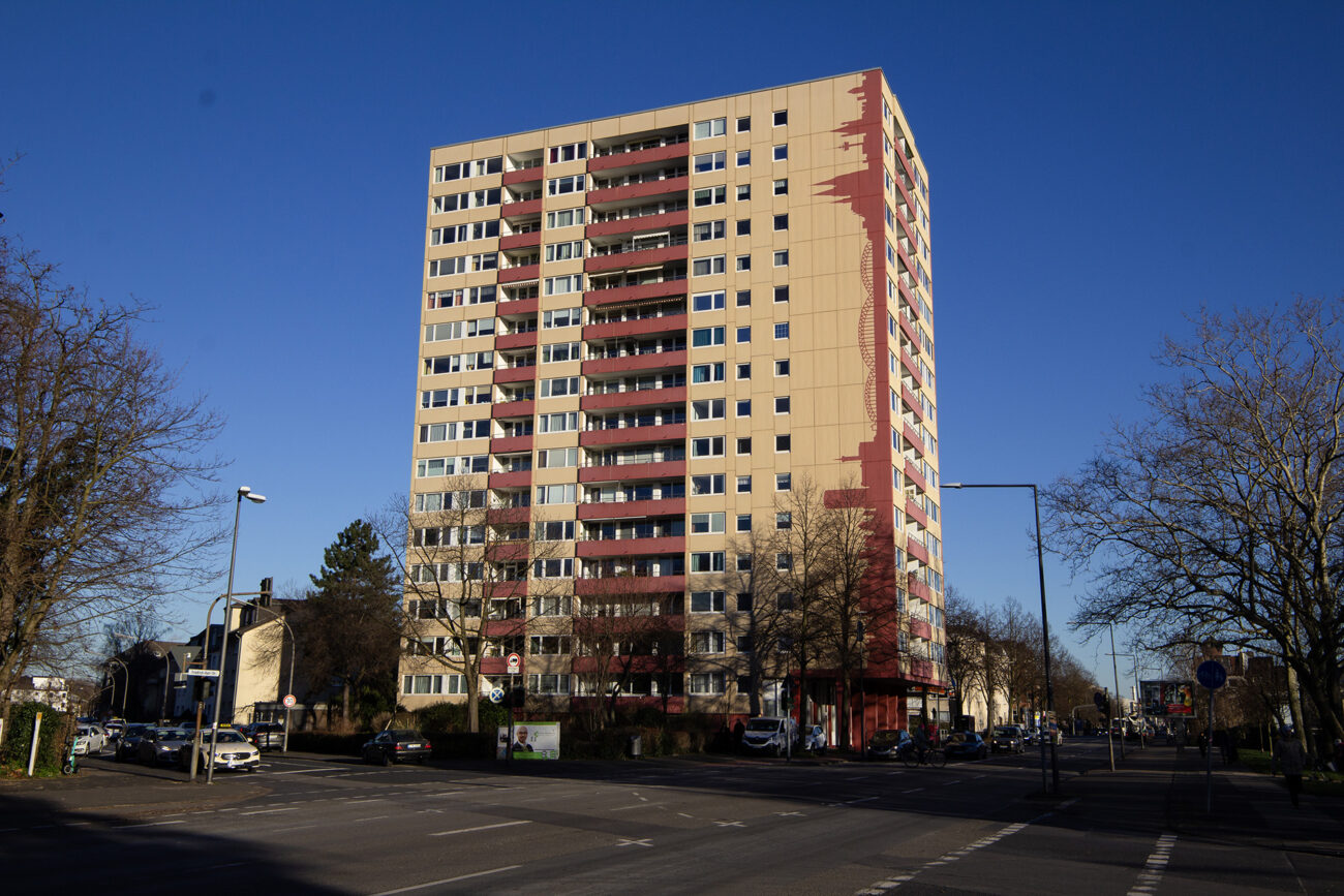 High-rise building in Cologne-Niehl featuring a graphic mural by Goodlack Art, in yellow and red, modern urban art, sunny day, blue sky, urban setting with cars and trees.