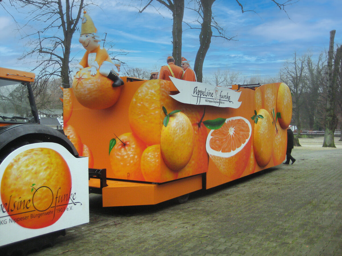 A company float with a graffiti design in Cologne, painted with bright orange oranges and a cartoon character in a carnival costume sitting on an orange. The float is part of a carnival parade and features the "Appelsine Funke" logo of the KKG Nippeser Bürgerwehr 1903 e.V.
