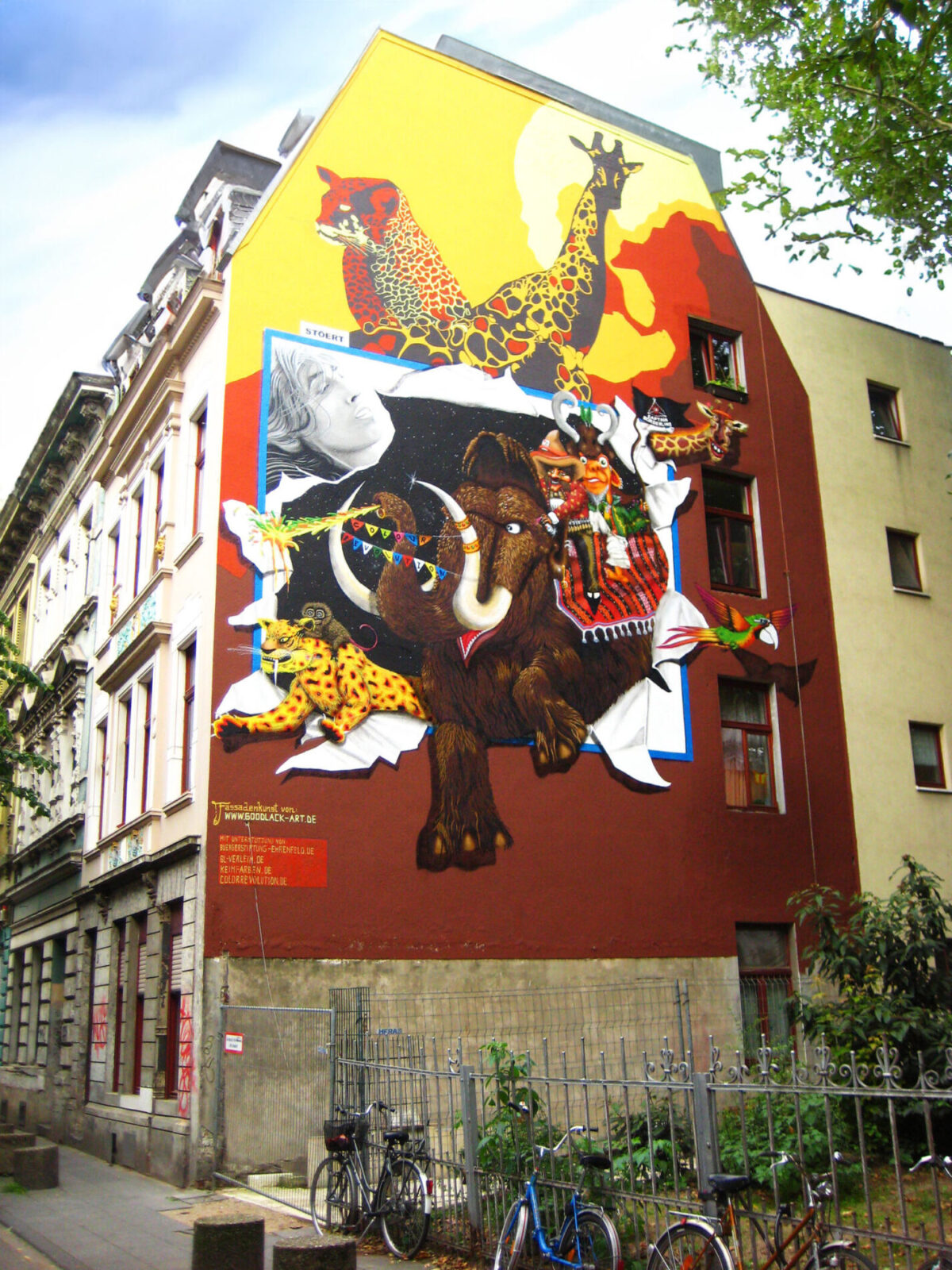Street Art in Cologne - An impressive mural by Goodlack Art features colorful animals such as leopards, giraffes, and a mammoth. The artwork spans an entire building wall, blending vibrant colors and dynamic motifs to create a striking urban landscape.