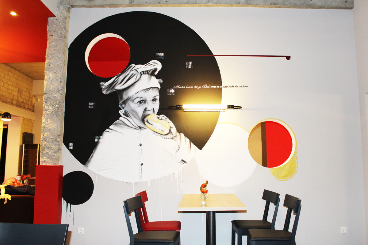Wandgemälde in einem Restaurant zeigt ein Kind, das in ein Brötchen beißt. Die Kunst kombiniert Schwarz-Weiß-Illustrationen mit kräftigen roten und gelben geometrischen Formen. Urban Streetart von Goodlack Art, inte