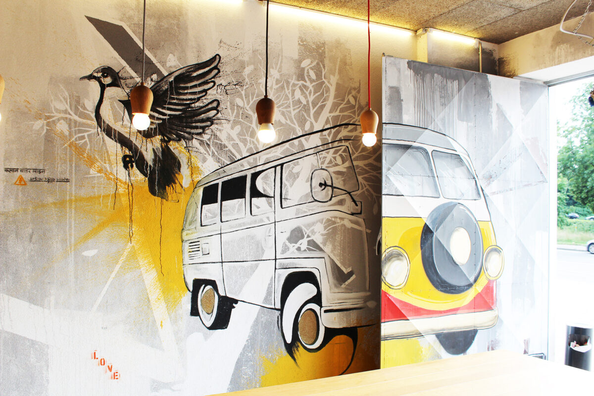 Streetart und Urbanart von Künstler Goodlack Art, zeigt eine Wandmalerei mit einem stilisierten Vogel und einem Retro-Bus in gelben und grauen Tönen, beleuchtet von modernen Lampen. Kunstgrafik mit urbanem Flair.