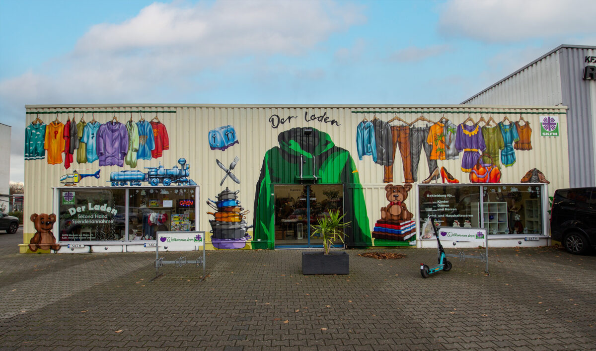 Buntes Graffiti an der Fassade eines Second-Hand-Ladens in Monheim, das Kleidung, Spielzeug und Haushaltswaren darstellt. Im Vordergrund steht ein Schild mit der Aufschrift 'Der Laden'. Professionelle Fassa