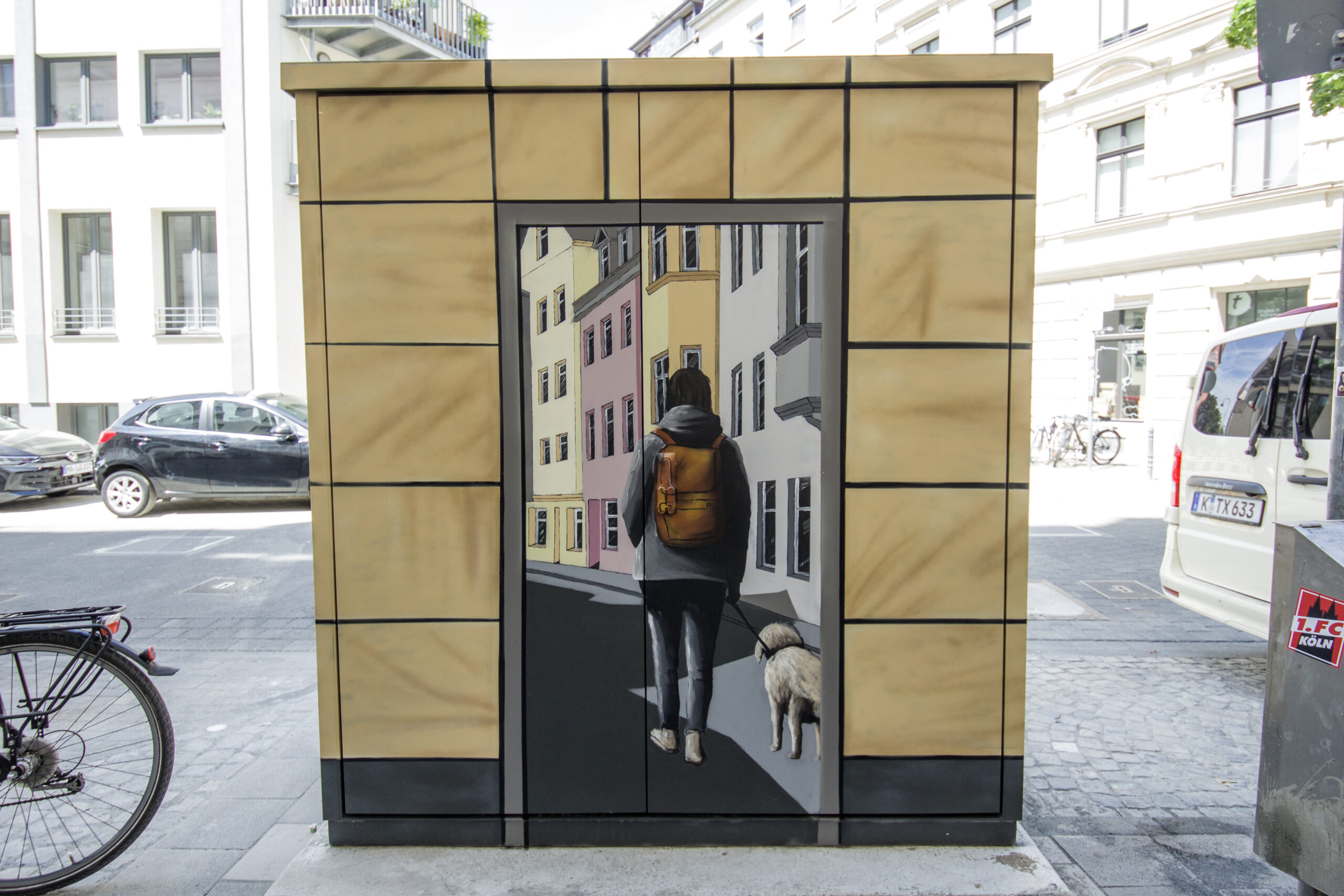 Stra&szlig;enkunst auf einem Verteilerkasten in K&ouml;ln, zeigt eine realistisch gemalte Szene einer Person mit Rucksack, die mit einem Hund eine Stra&szlig;e entlanggeht. Die Szene ist von Geb&auml;udezeichnungen eingerahmt, die den Kasten k&uuml;nstlerisch gestalten. Im Hintergrund sind parkende Autos und Geb&auml;ude zu sehen, typisch f&uuml;r urbane Street Art.